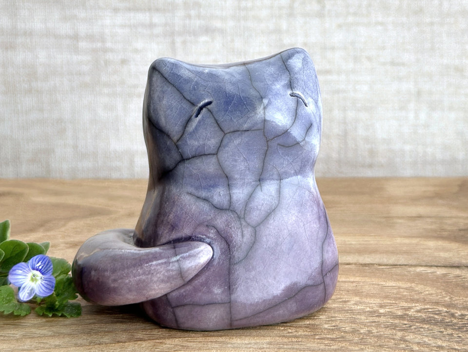 A calm and gentle Shinto Inari nighttime kitsune fox spirit ceramic raku statue glazed in a bottom-to-top gradient of purple to blue. Its tail is curled around the right side of its body, and it has a kind face.