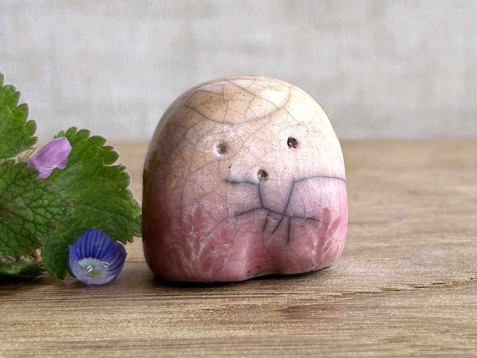 a small and stout nature spirit raku sculpture glazed in peach. it has a cute little face, and little plant carvings on its front. there are tiny dark grey spiderweb patterns in the glaze, in addition to the normal raku crackles, from where raindrops fell on it.