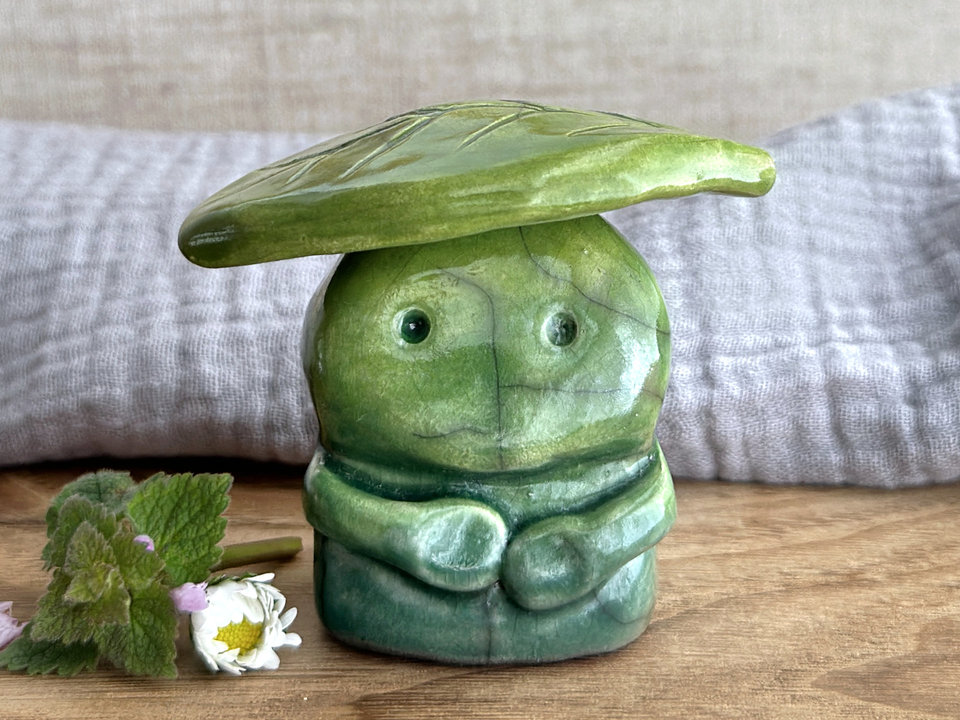 Raku ceramic sculpture glazed in forest green to spring green (bottom to top) with a kind, but slightly intense, face, and a brighter yellow-green leaf on its head. It also has two cute little arms.