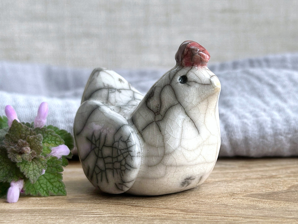 a white raku ceramic chicken with grey eyes, a pale orange beak, and a bordeaux red comb on the top of her head. she looks very sweet. There are tiny dark grey spiderweb patterns in the glaze, in addition to the normal raku crackles, from where raindrops fell on it.