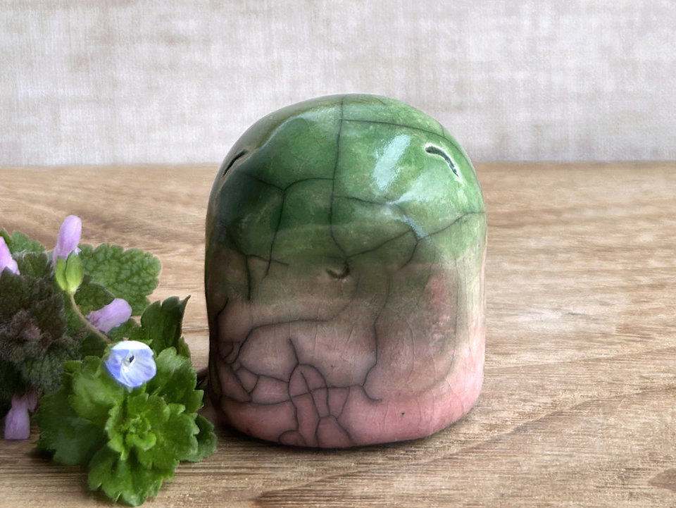 raku kodama kami nature spirit sculpture glazed in a bottom to top gradient of soft reddish pink to spring green. it has a gentle, kind face. there are tiny dark grey spiderweb patterns in the glaze, in addition to the normal raku crackles, from where raindrops fell on it.