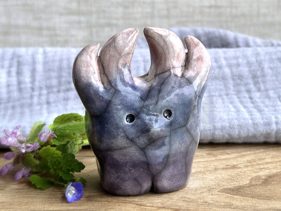 a raku ceramic creature. it has four stubby legs and gets wider as its body goes up. it eventually ends in four small antlers. on the way, it has a gentle face with a cute little nose. it's glaze in a bottom-to-top gradient of dark purple to grey-blue to a pale red-pink for the antlers. it is a friend.