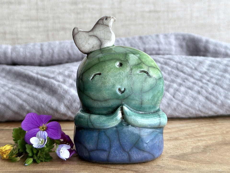 a raku ceramic jizo nature spirit guardian sculpture glazed in a bottom-to-top gradient of blue to forest green to spring green. they have a happy, smiley face, little praying hands, and a sweet white bird on their head.