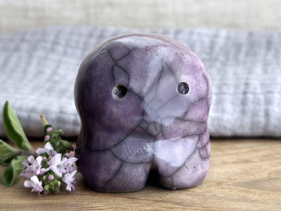 A kodama nature spirit ceramic guardian statue glazed in deeper and lighter purple-violet colours. It has four stubby legs, a wide face, and two big eyes.