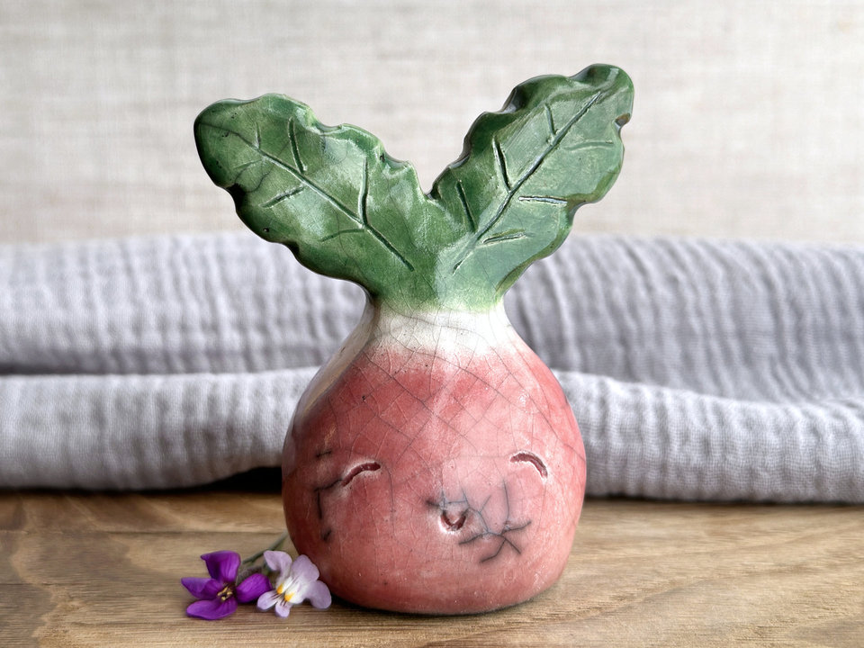 a very smiley and happy radish spirit raku ceramic sculpture. it has a round body glazed in a soft radish red colour. the top of its body fades to white, and it has two green leaves on the top of its head/body.