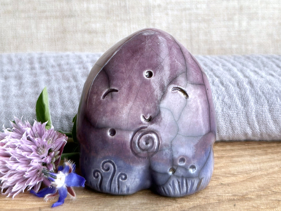 round and vaguely egg-shaped nature spirit sculpture. it is glazed in a top-to-bottom gradient of purple to indigo violet and has spiral carvings on its body. it also has a gentle, happy face.