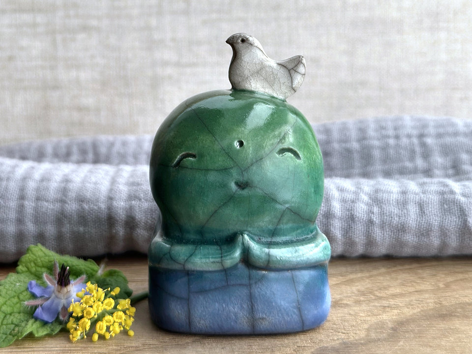 a raku ceramic jizo nature spirit guardian sculpture glazed in a bottom-to-top gradient of blue to forest green to spring green. they have a happy, smiley face, little praying hands, and a sweet white bird on their head.