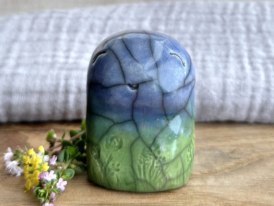 an earth elemental nature spirit ceramic sculpture. it is glazed in a bottom-to-top gradient of forest and spring green to blue and has plant and flower carvings on its body. it has a gentle, smiling face.