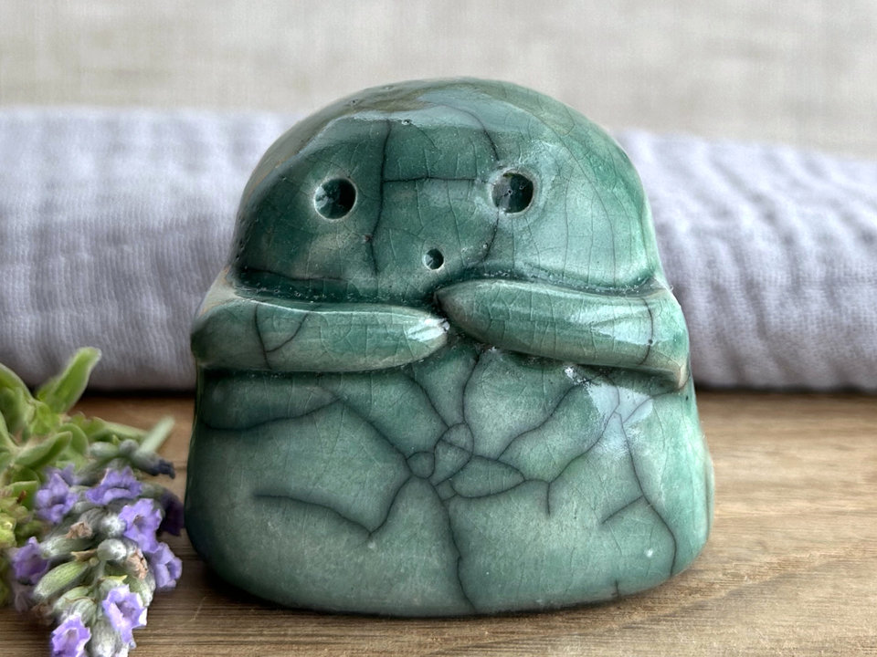A sweet little kodama nature spirit ceramic guardian statue glazed in soft forest green and with a lovely, gentle face and little arms. they have some very small, dimpled, pinhole glaze disturbance marks on their back and side.