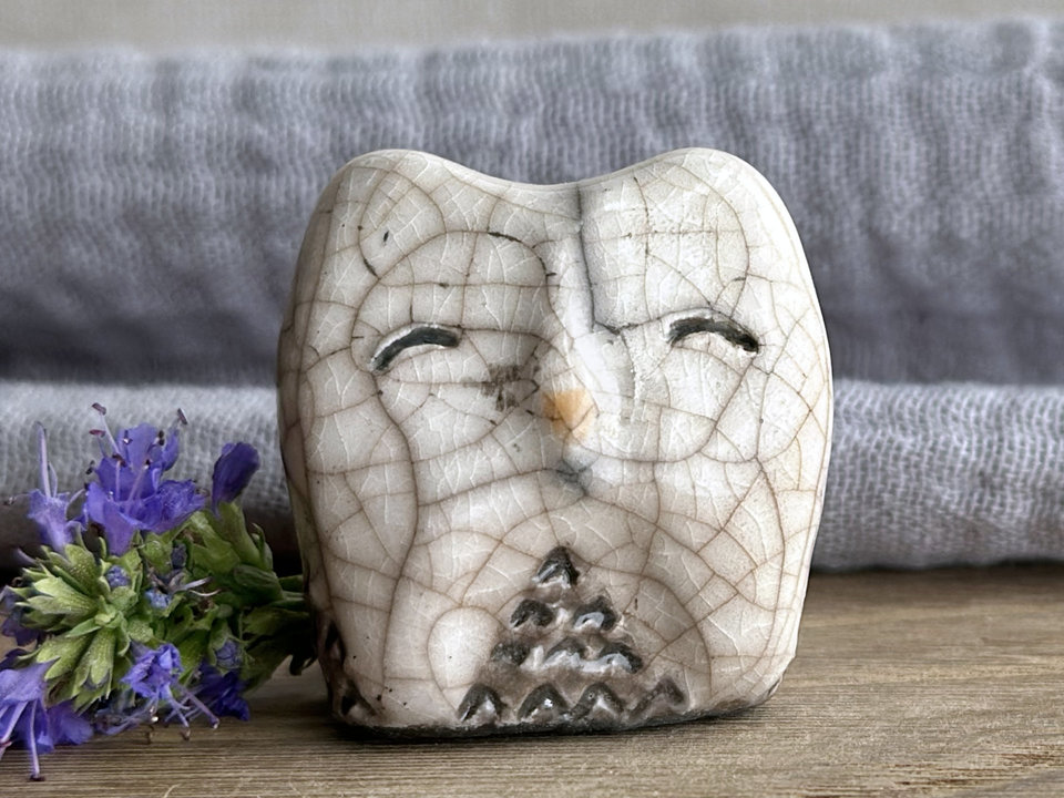 Raku ceramic little owl or owlet kami nature spirit statue with sort of pointed ears. It’s glazed in white and grey-brown with gentle, smiling eyes and a little golden yellow beak.