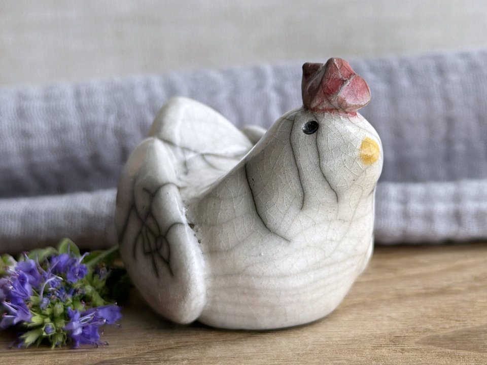 a white raku ceramic chicken with grey eyes, a pale orange beak, and a bordeaux red comb on the top of her head. she looks very sweet.