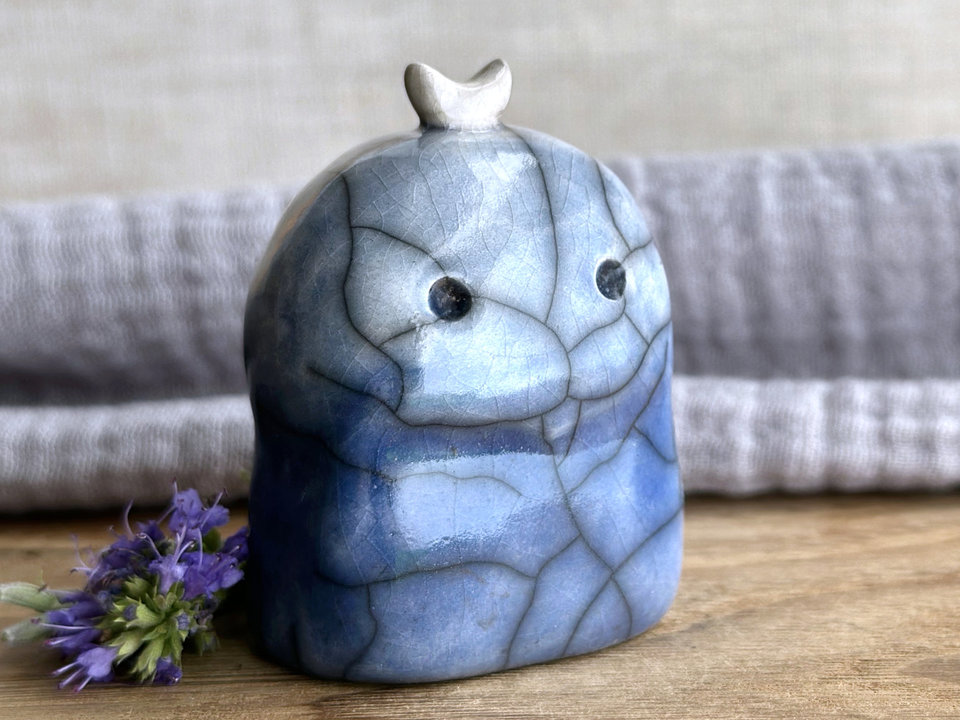 A kodama nature and forest spirit kami ceramic raku sculpture glazed in a bottom-to-top gradient of darker to lighter blue. It has a gentle-but-worried face, and a white crescent moon on its head.