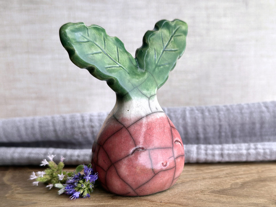 a very smiley and happy radish spirit raku ceramic sculpture. it has a round body glazed in a soft radish red colour. the top of its body fades to white, and it has two green leaves on the top of its head/body.