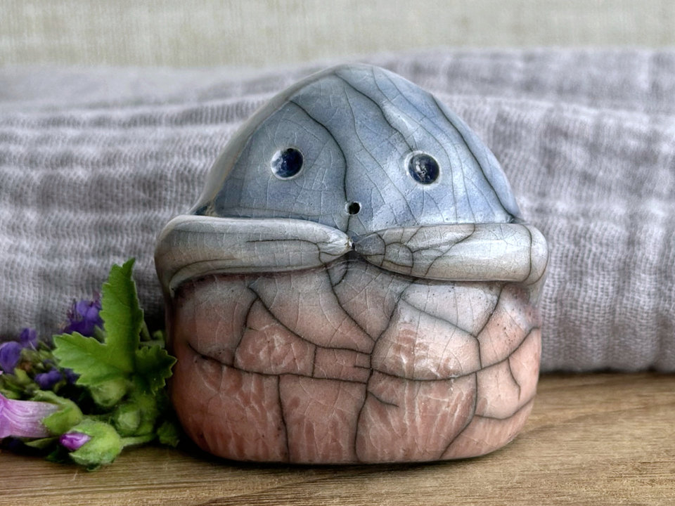 a vaguely dome-shaped nature spirit ceramic sculpture. it is glazed in a bottom-to-top gradient of soft, pale reddish pink to pale blue and has plant and flower carvings on its body. it also has a gentle face, and two little arms across its front.