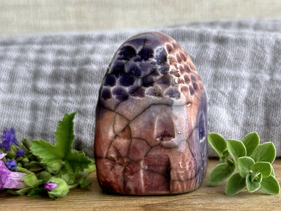 Tiny raku ceramic cottage glazed in warm red-orange, bordeaux red, mauve, and purple. Its roof is carved to look like slates. It has a carved door and windows and lots of plant and flower carvings on its walls.