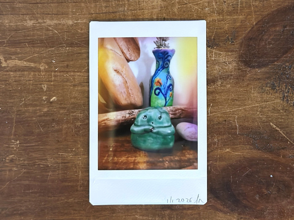 an instax mini print of a small, forest green raku ceramic kodama with a gentle face and two cute little arms. it's on a wooden shelf, and there are various pieces of wood and also a small bottle painted with plant designs in blue, green, purple and orange behind it.