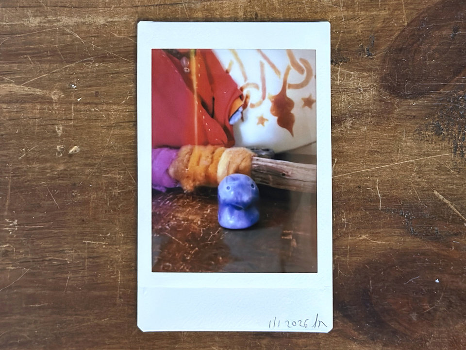 an instax mini print of a small violet nature spirit sculpture with a gentle face. it's standing on a wooden shelf, and behind it is a stick wrapped in plant-dyed wool in warm pink and orange. behind that are some red cloth and the bottom of a traditional drum decorated with henna.
