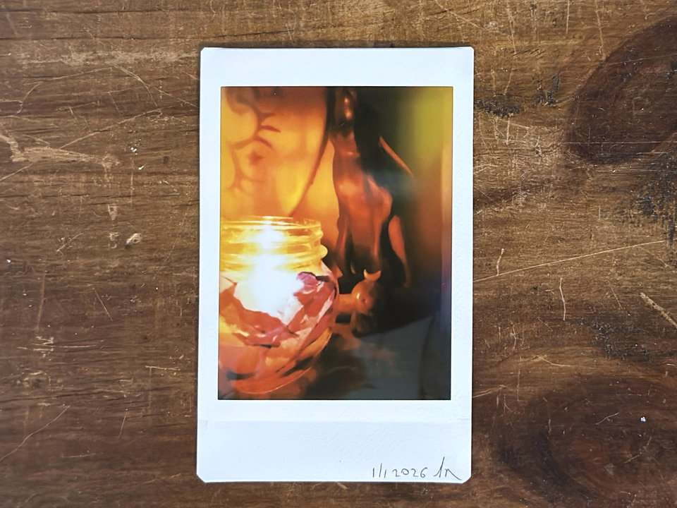 a dark, atmospheric instax mini print of a blue raku ceramic kodama guardian nature spirit with a white crescent moon on their head. the scene is lit by a candle in a glass jar that's covered in tissue paper in autumnal reds, oranges, golden yellows, and purples. there's a wooden hare in the background along with the edge of a traditional drum.