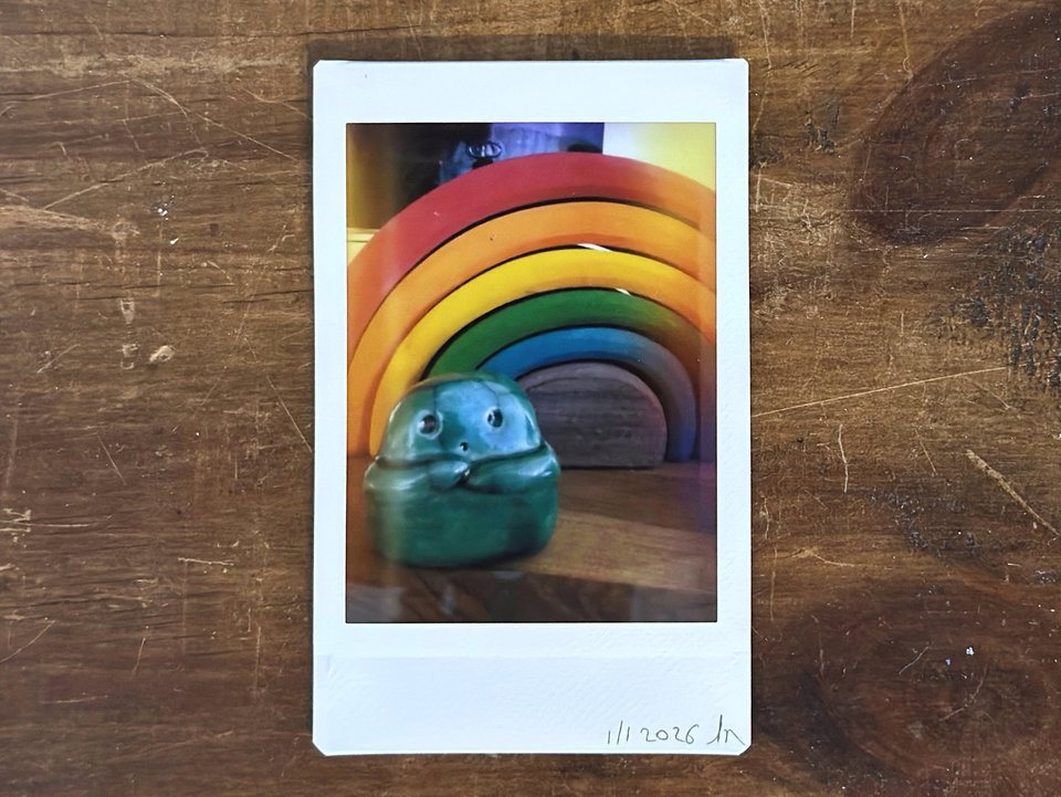 an instax mini print of a small, forest green raku ceramic kodama with a gentle face and two cute little arms. it's on a wooden shelf, and there's a stacking wooden rainbow behind it.