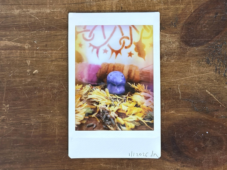 an instax mini print of a small, violet guardian nature spirit. there are lots of dried, pale golden orange calendula flowers and lavender flowers around them. behind them is a stick wrapped in plant-dyed wool in warm pink and orange, and behind that is the bottom of a traditional drum decorated with henna.