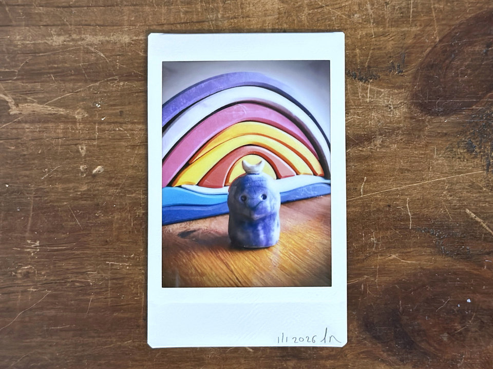 an instax mini print of a blue raku ceramic kodama guardian nature spirit with a white crescent moon on their head. they're standing on a piece of wood, and there's a wooden stacking shape behind them that looks like sunset on the water. the two base pieces are blue, and there's then a rainbow shape above them with reds, oranges, yellows, pinks, and purples in it.