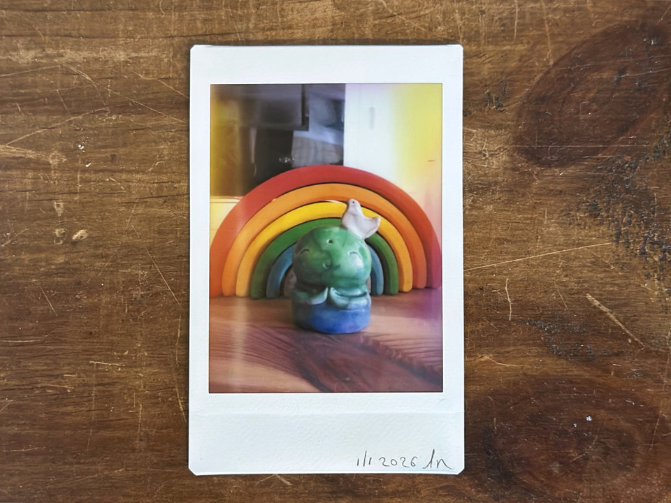 an instax mini print of a raku ceramic jizo nature spirit guardian sculpture glazed in a bottom-to-top gradient of blue to forest green to spring green. they have a happy, smiley face, little praying hands, and a sweet white bird on their head. they're standing in front of a wooden stacking rainbow.