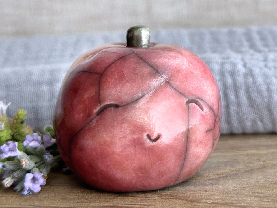 raku ceramic warm-red apple nature spirit sculpture with a happy, gently smiling face. it has a cute little green-brown stalk on top.