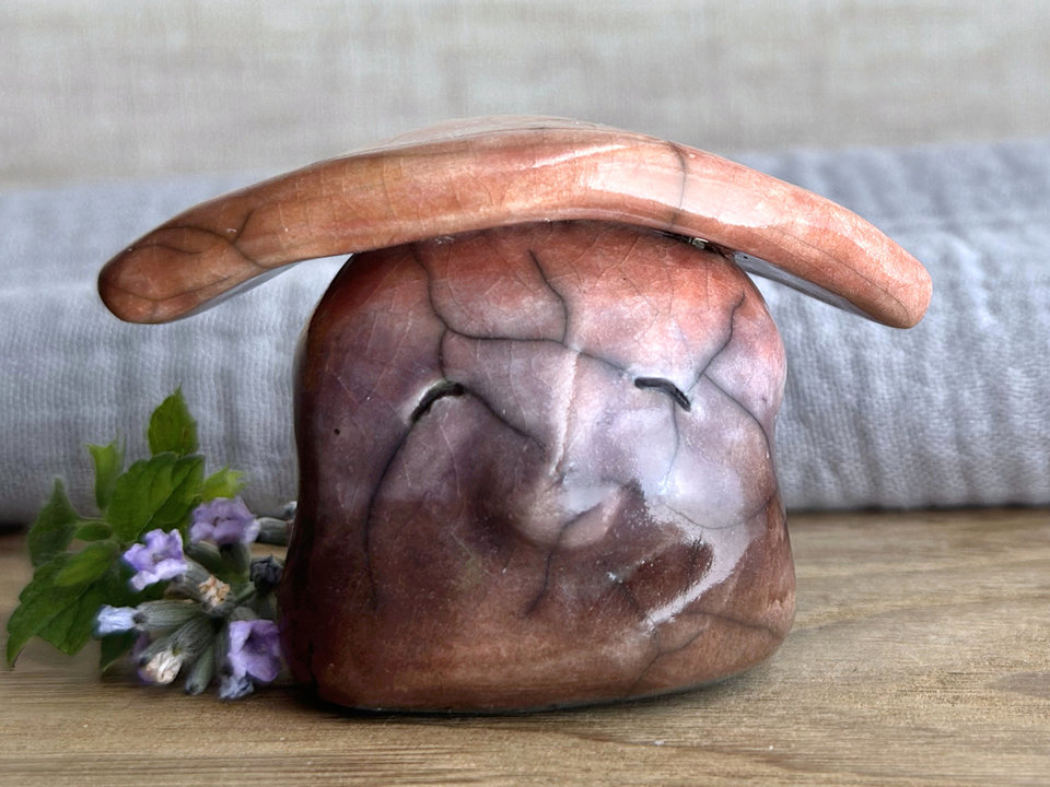 Raku ceramic sculpture glazed in a bottom-to-top gradient of brown to violet to mauve to warm orange with a kind and gentle face, and a warm-orange and golden yellow leaf on its head.