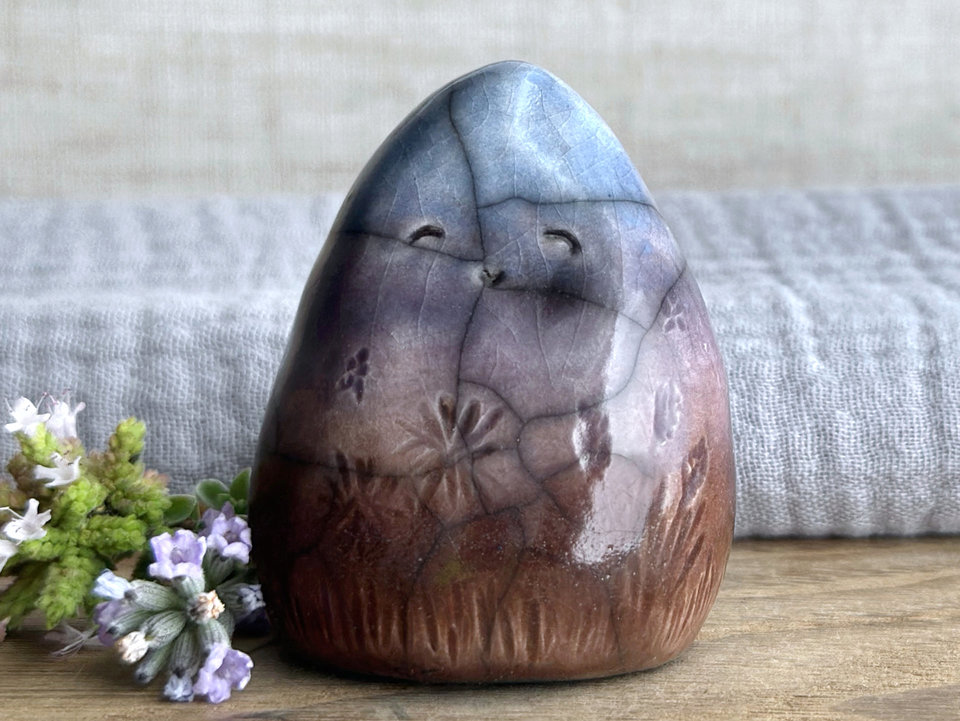 vaguely dome-shaped nature spirit ceramic sculpture. it is glazed in a bottom-to-top gradient of a brown to soft mauve to violet to blue and has autumnal plant and flower carvings on its body. it has a gentle, smiling face.
