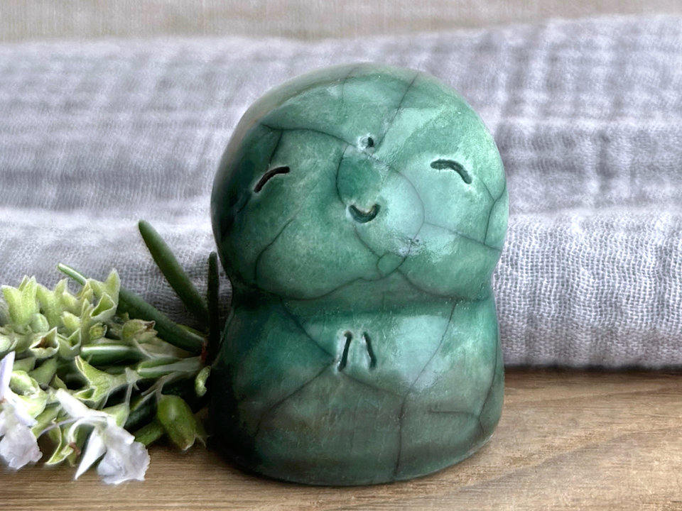 a smiley and loving Jizo Shinto raku ceramic sculpture talisman glazed in forest green with a hint of brown at the bottom. they have a kind, gentle face and are a wonderful smooth shape to hold in the hand.