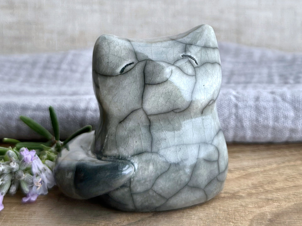 A Shinto Inari kitsune fox spirit ceramic raku statue glazed in grey. Its tail is curled around the right side of its body and has a darker grey tip. It has a kind face.