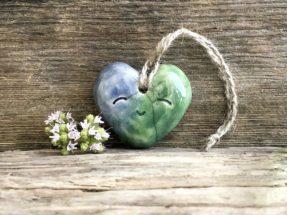 A smiley blue and green heart ornament in raku ceramic. It has a hole in the top, and there is a piece of twine threaded through the hole to act as a hanger.