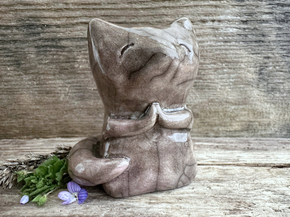 Raku ceramic fox sculpture with a kind, gentle face and sweet praying hands. It is glazed in a warm brown colour with a hint of soft pink, and its tail is curled around its body.