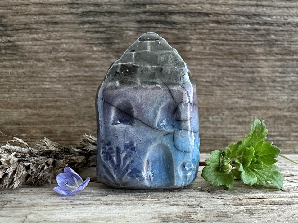 Tiny raku ceramic cottage glazed in blue, violet, and turquoise. Its roof is glazed in grey and carved to look like slates. It has a carved door and windows and lots of plant and flower carvings too.