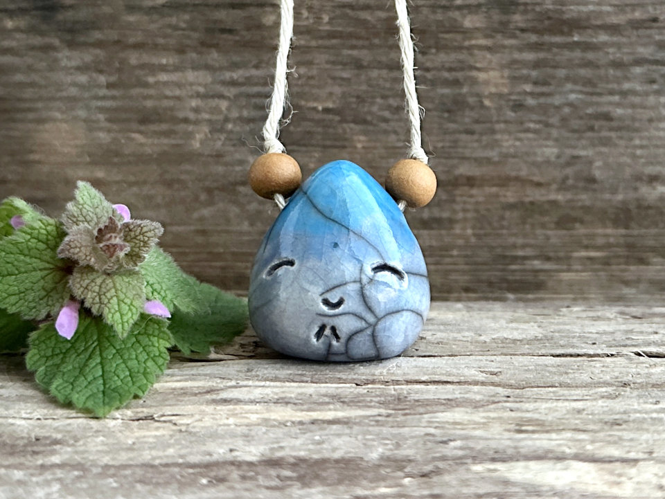raku ceramic pendant in the shape of a tiny raindrop with a happy face. it's glazed in a gradient from blue to aqua-turquoise (bottom to top) and strung on an eco flax cord. there is a round sandalwood bead on either side of the pendant.