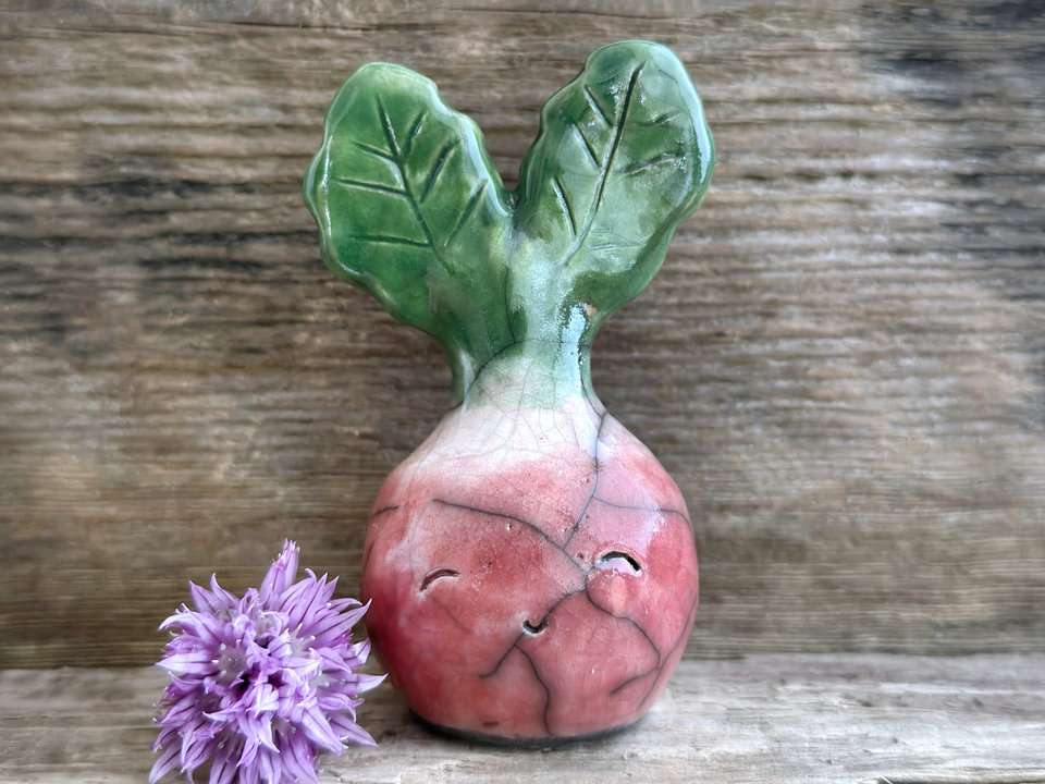 a very smiley and happy radish spirit raku ceramic sculpture. it has a round body glazed in a soft radish red colour. the top of its body fades to white, and it has two green leaves on the top of its head. body. it is round with green leaves on top!