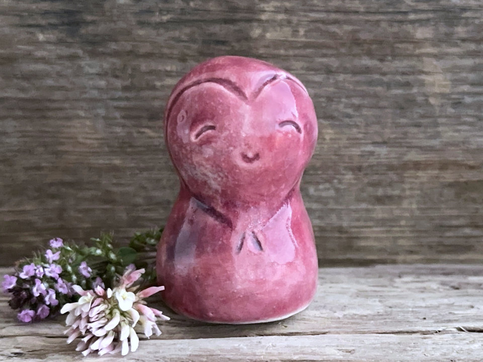 A smiley and loving Jizo Shinto raku ceramic sculpture talisman glazed in warm, pinky red colour with hints of purple. It has a heart shape carved around its kind, gentle face and is a wonderful smooth shape to hold in the hand.