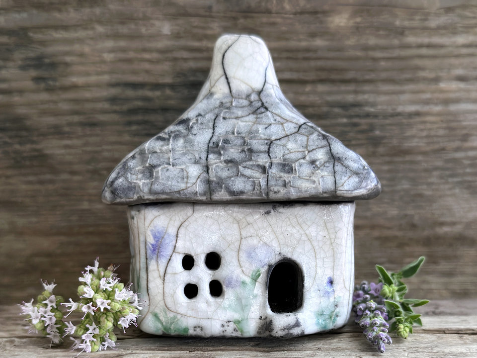 Rustic raku ceramic kurinuki cottage glazed in white with pale green plant and pale violet flower patterns on the walls. The roof has a chimney and is glazed in grey with hints of white to look like slate. It has a cute little doorway and lots of windows.
