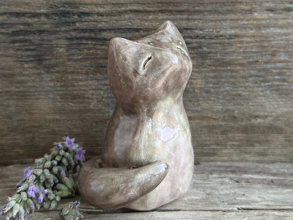 a beautiful Shinto Inari kitsune fox kami spirit ceramic raku statue. it's looking up with a peaceful, happy expression on its gentle face and is glazed in soft browns and pink-browns with its tummy being lighter than the rest. its tail is curled around its body on its right side, and it has white, snow-like marks in the glaze on the left side of its head and back.