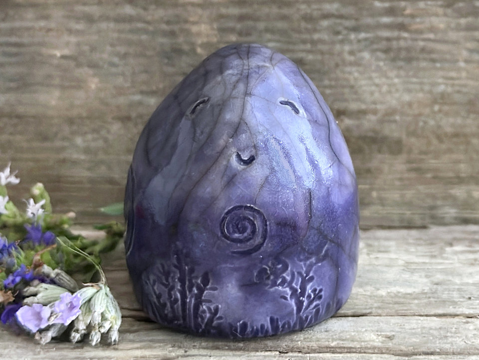vaguely egg-shaped gnome nature spirit sculpture. it is glazed in a bottom-to-top gradient of darker to lighter violet and has spiral and plant carvings on its body. it also has a gentle, happy face and a cute little nose.