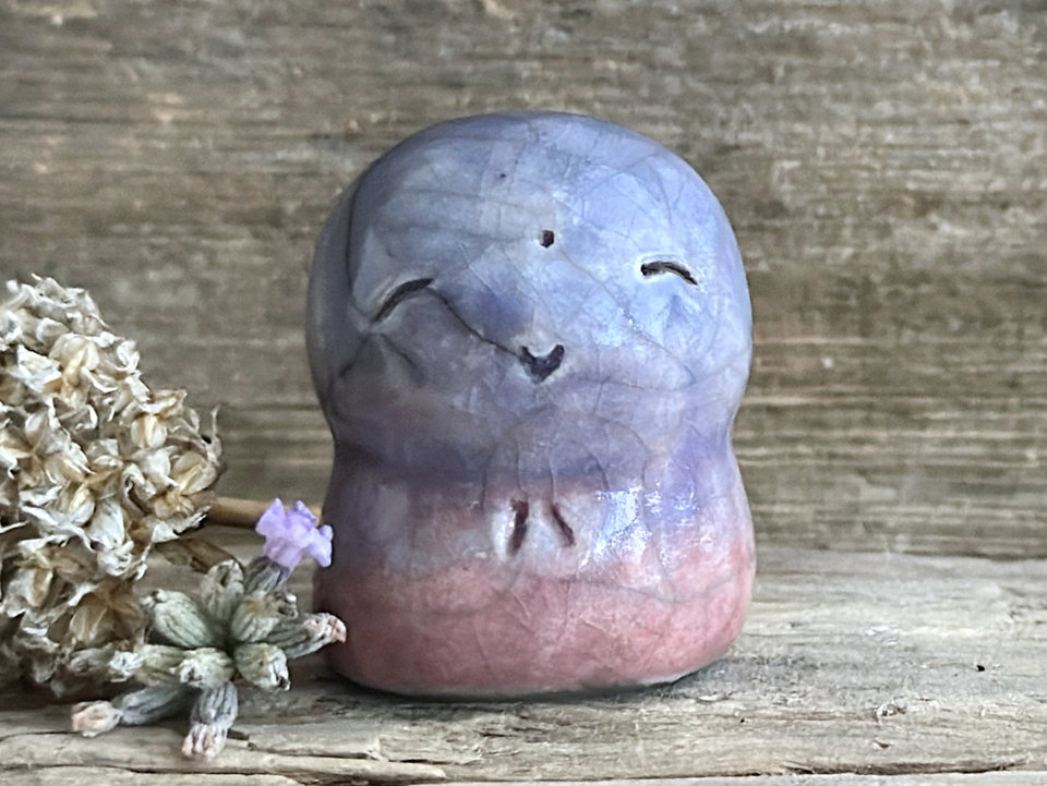 A smiley and loving Jizo Shinto raku ceramic sculpture talisman glazed in a bottom-to-top gradient of soft mauve to purple to blue. It has a kind, gentle face and is a wonderful smooth shape to hold in the hand.