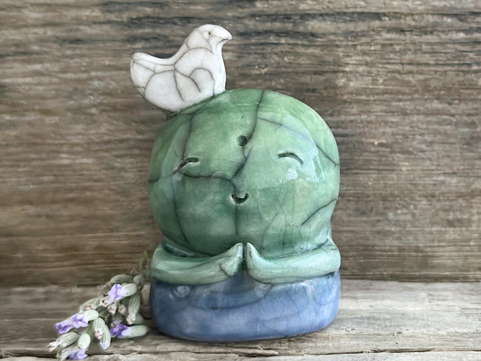 a shinto jizo nature spirit guardian raku ceramic sculpture glazed in a bottom-to-top gradient of blue to forest green to spring green. they have a happy, smiley face, little praying hands, and a sweet white bird on their head.