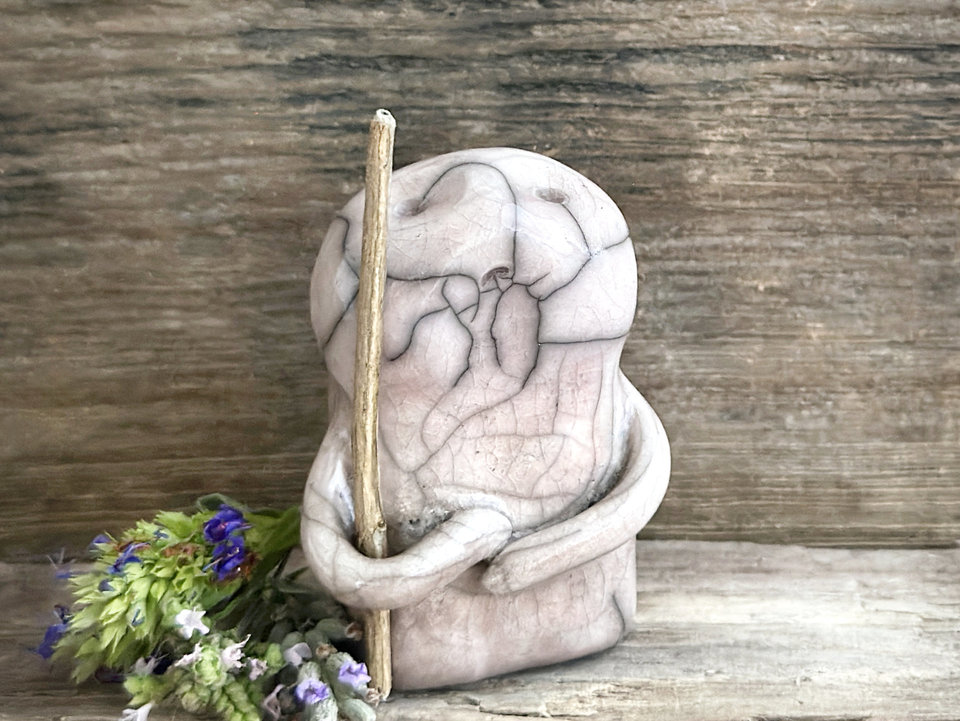 raku ceramic nature spirit warrior sculpture. it is glazed in very pale, soft mauve colour and carries a brown stick in one of its arms. it has a sad and worried but determined look on its gentle face.