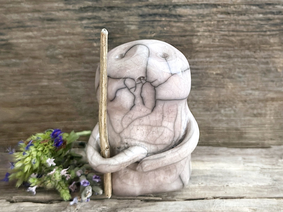 raku ceramic nature spirit warrior sculpture. it is glazed in very pale, soft mauve colour and carries a brown stick in one of its arms. it has a sad and worried but determined look on its gentle face.