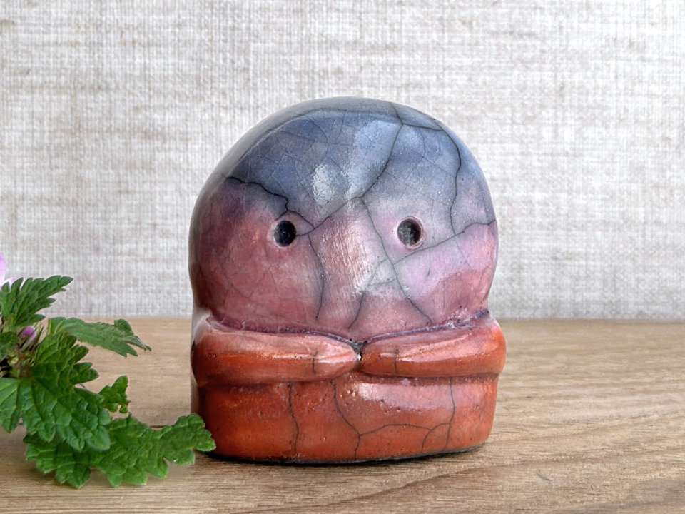 A kodama nature spirit ceramic guardian statue glazed in a top-to-bottom gradient of blue to violet to mauve to red-orange. It has a wide face, two big eyes, and cute little arms.