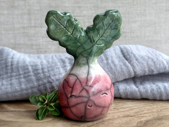a very smiley and happy radish spirit raku ceramic sculpture. it has a round body glazed in a soft radish red colour. the top of its body fades to white, and it has two green leaves on the top of its head. body. it is round with green leaves on top!