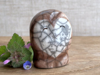 Ceramic raku owl kami nature spirit sculpture glazed mostly in soft brown with a white heart-shaped face. It has a very kind, friendly expression. There are tiny dark grey spiderweb patterns in the glaze, in addition to the normal raku crackles, from where raindrops fell on it.