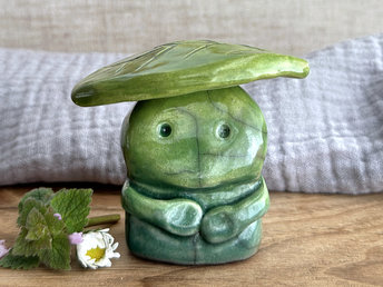 Raku ceramic sculpture glazed in forest green to spring green (bottom to top) with a kind, but slightly intense, face, and a brighter yellow-green leaf on its head. It also has two cute little arms.