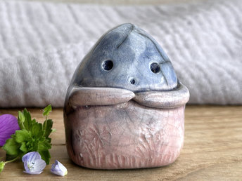 a vaguely dome-shaped nature spirit ceramic sculpture. it is glazed in a bottom-to-top gradient of soft reddish pink to blue and has plant and flower carvings on its body. it also has a gentle face, and two little arms across its front.
