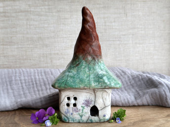 a rustic raku ceramic kurinuki cottage glazed in white with green plant carvings and purple, violet, and indigo flower carvings on the walls. the roof has a very tall, pointy and slightly twisty chimney and is glazed in brown on the pointy bit and shades of green on the lower bit to look a bit like a tree. the cottage has a cute little doorway and lots of windows.